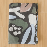 A folded length of cotton linen blend fabric, featuring an abstract painterly pattern in olive green, black, white, light blue, and dusty pink, laid flat on a light wood background.