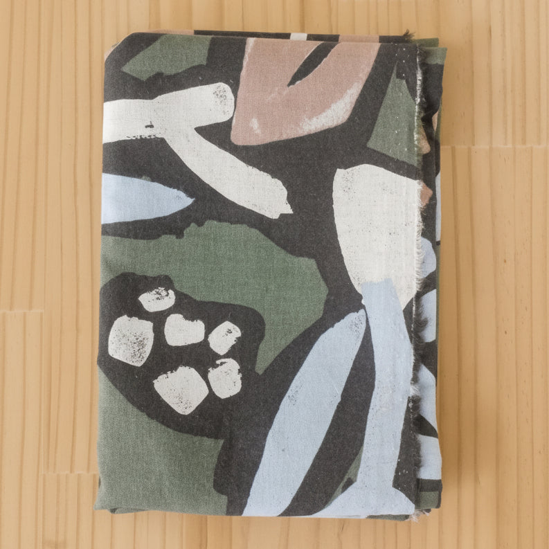 A folded length of cotton linen blend fabric, featuring an abstract painterly pattern in olive green, black, white, light blue, and dusty pink, laid flat on a light wood background.