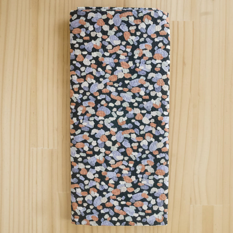 Folded fabric end of bolt remnant, featuring a dense multicolor terrazzo pattern on a black background, laid flat on a light wood surface.