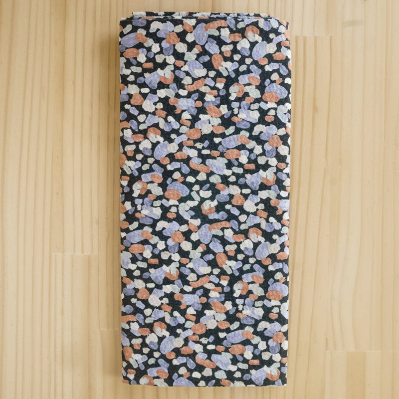 Folded fabric end of bolt remnant, featuring a dense multicolor terrazzo pattern on a black background, laid flat on a light wood surface.