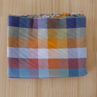 A folded plaid organic cotton fabric length with selvedge showing, featuring a multicolor check pattern in blue, orange, red, beige, and white, laid flat on a light wood background.