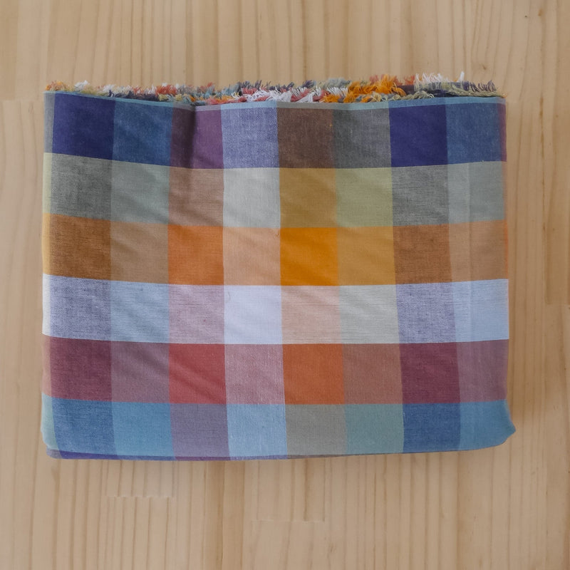 A folded plaid organic cotton fabric length with selvedge showing, featuring a multicolor check pattern in blue, orange, red, beige, and white, laid flat on a light wood background.