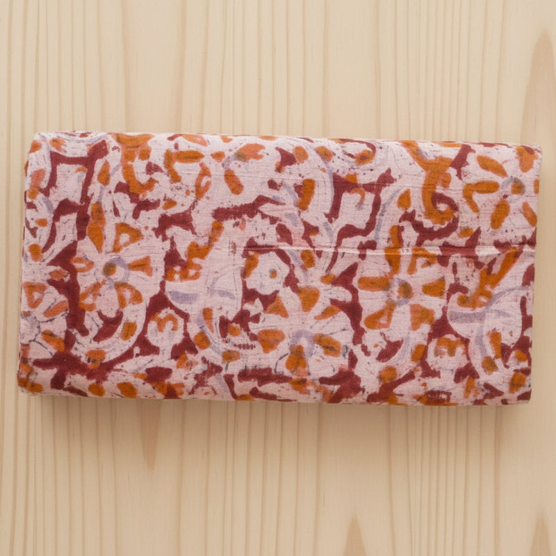 A folded rectangular block print Indian cotton fabric, featuring an abstract floral block-print pattern in orange, dark red, and light purple on a pale pink base, laid flat on a light wood background.