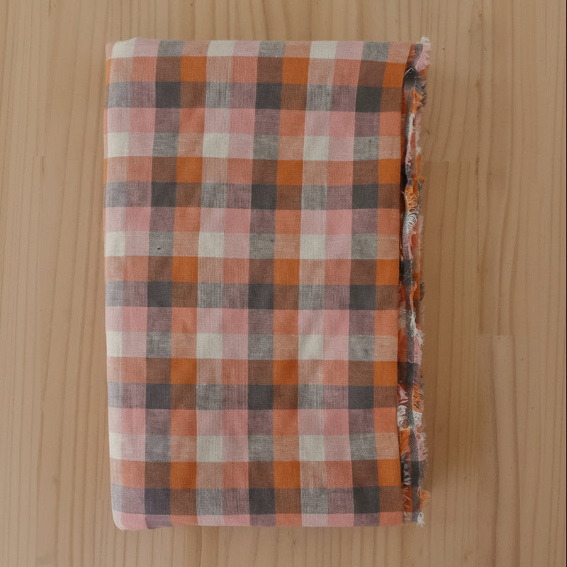 A folded piece of cotton linen blend plaid fabric with orange, pink, white, and gray checkered stripes, displayed flat on a light wood grain surface.