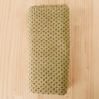 Folded cotton fabric in a light olive green with a darker olive green cross stitch thread woven over the surface of the fabric, laid flat on a light wood grain surface.