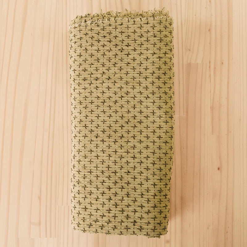 Folded cotton fabric in a light olive green with a darker olive green cross stitch thread woven over the surface of the fabric, laid flat on a light wood grain surface.