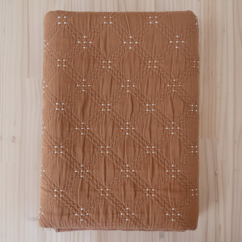 A folded brown quilted cotton jacquard fabric with a diamond pattern and small white embroidered details, laid flat on a light wood floor.