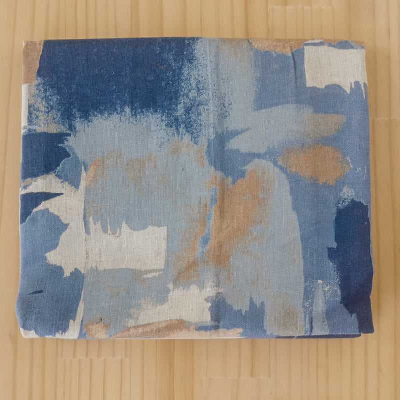 A length of folded linen cotton blend fabric with an abstract, painterly design in shades of blue, beige, and white, displayed flat on a light wood background.