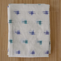 A folded white cotton Ikat fabric with a repeating pattern of small blue and green dash marks, displayed on a light wood background.
