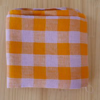 A square folded linen fabric item, featuring a gingham checkered pattern in orange and light lavender, displayed on a light wood grain surface.
