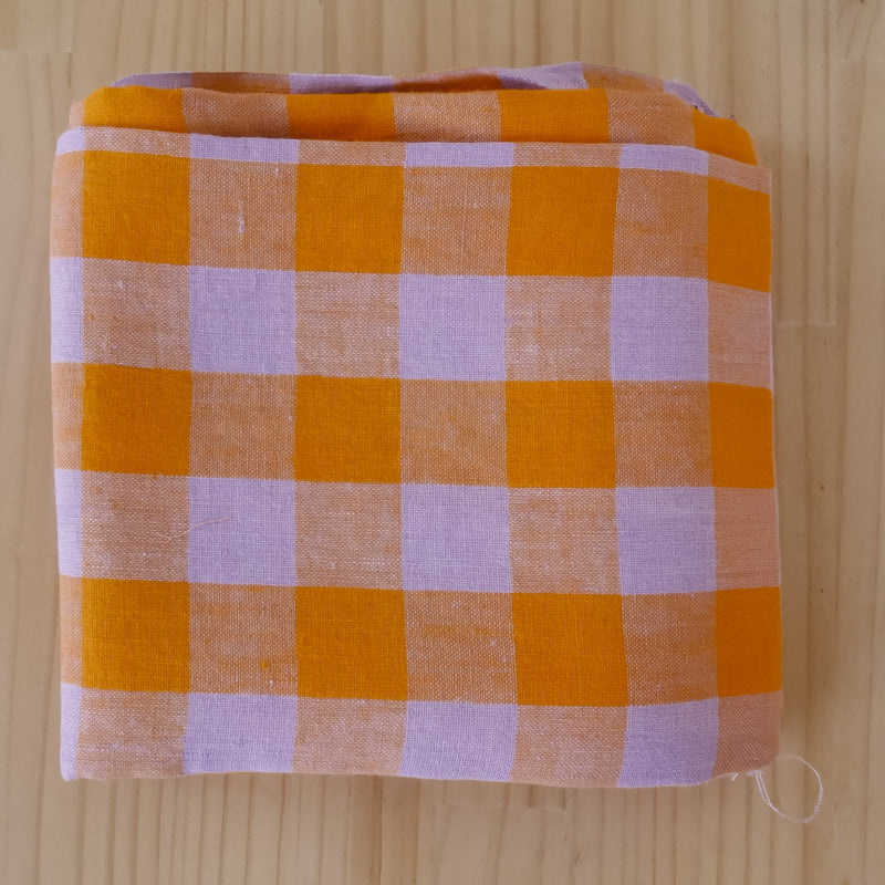 A square folded linen fabric item, featuring a gingham checkered pattern in orange and light lavender, displayed on a light wood grain surface.
