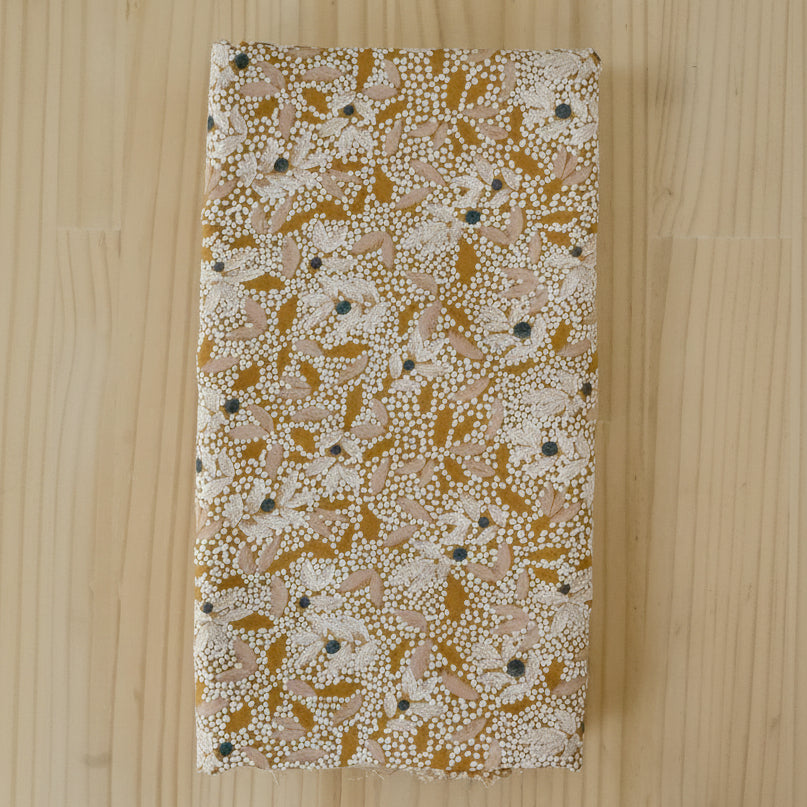 A folded length of viscose crepe fabric with an abstract floral print in white, mustard and pale pink on a light wooden background.