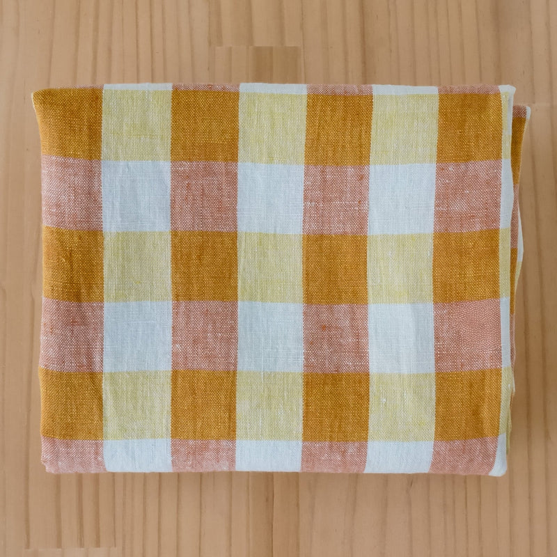 A folded piece of linen fabric with a gingham check pattern in mustard yellow, dusty pink, pale yellow, and white, laid flat on a light wood background.