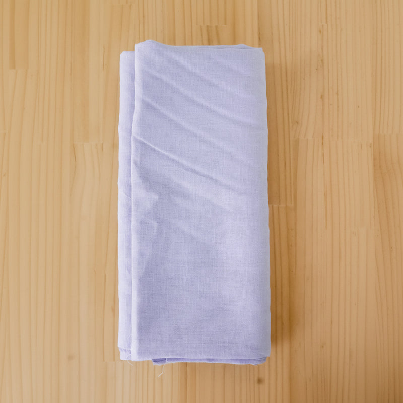 A folded piece of light lavender linen fabric, displayed on a light wood grain surface.