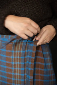 Woman wearing a blue plaid Nevis skirt with a dark brown knit top and boots, highlighting the tie waist detail of the skirt. The Nevis Skirt Sewing Pattern is designed by Merchant and Mills and available at Stitch Piece Loop Australia.